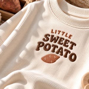以下が含まれることがあります： クリーム色のリブ編みの長袖シャツで、茶色の刺繍で「LITTLE SWEET POTATO」という文字が施されています。その文字のすぐ下には、茶色の刺繍で描かれた小さなサツマイモのグラフィックが胸元に配置されています。