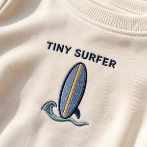 Puede incluir: Una sudadera de color crema con un diseño bordado de una tabla de surf azul con una franja central amarilla y una ola verde azulada. El texto "TINY SURFER" está bordado en letras mayúsculas azul oscuro sobre el gráfico.