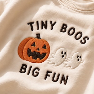 May include: A cream-coloured sweatshirt featuring dark brown embroidered text that reads TINY BOOS BIG FUN. The design includes an orange jack-o'-lantern pumpkin with a dark brown face and two small white embroidered ghosts with black eyes.