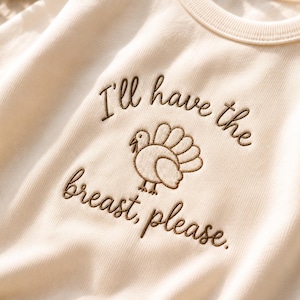 Könnte beinhalten: Ein cremefarbenes Sweatshirt mit Rundhalsausschnitt und brauner Stickerei. Der Text "I'll have the breast, please." ist zusammen mit einer kleinen Truthahn-Grafik zwischen den beiden Textzeilen auf der Vorderseite eingestickt.