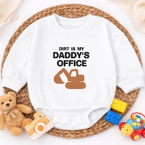 May include: White baby bodysuit with long sleeves. The text "DIRT IS MY DADDY'S OFFICE" is printed in black, with a brown excavator graphic. The bodysuit is surrounded by toys, including a teddy bear and building blocks.