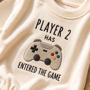 May include: A cream-colored long-sleeve sweatshirt featuring black embroidered text that reads PLAYER 2 HAS ENTERED THE GAME. Centered below the text is an embroidered graphic of a video game controller in beige with black and colorful buttons.