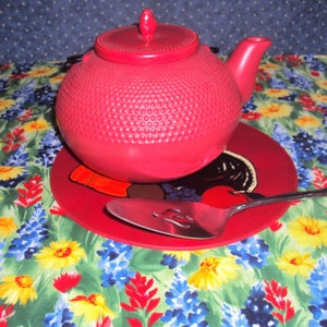 May include: A bright red teapot with a textured surface sits on a red and white floral patterned coaster. A silver serving spoon is on the coaster.
