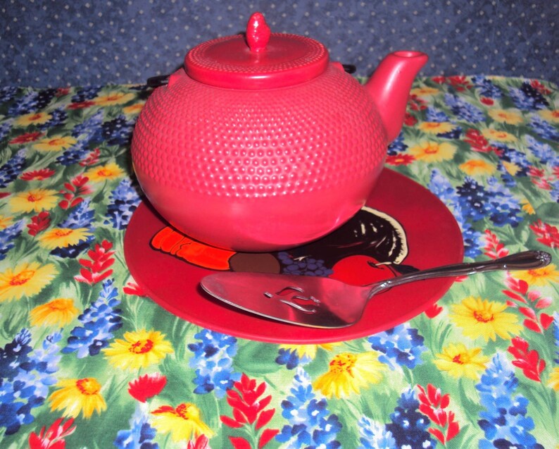 May include: A bright red teapot with a textured surface sits on a red and white floral patterned coaster. A silver serving spoon is on the coaster.