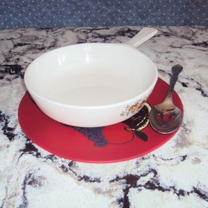 May include: A white ceramic bowl with a floral design sits on a red trivet with a fruit design. A silver slotted spoon is next to the bowl.