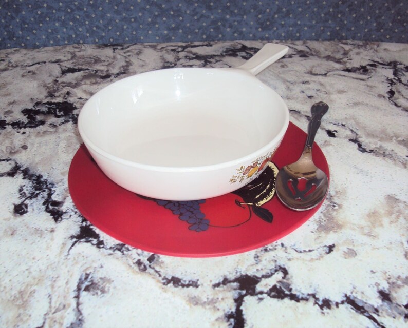 May include: A white ceramic bowl with a floral design sits on a red trivet with a fruit design. A silver slotted spoon is next to the bowl.