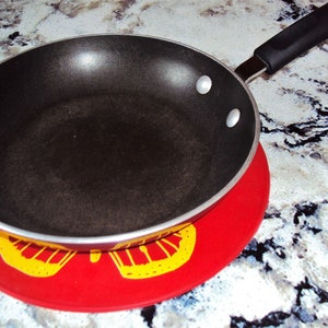 May include: A black non-stick frying pan with a silver handle sits on a red and yellow patterned hot pad.