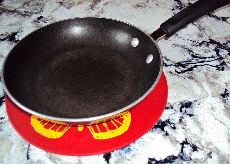 May include: A black non-stick frying pan with a silver handle sits on a red and yellow patterned hot pad.