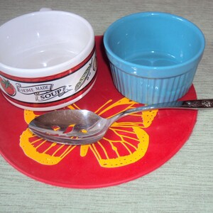 May include: A red butterfly-shaped silicone trivet with a white soup bowl with a green and red design that says "Home Made Soup" and a blue bowl. A silver slotted spoon is on top of the trivet.