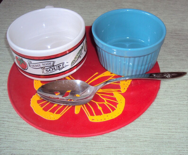 May include: A red butterfly-shaped silicone trivet with a white soup bowl with a green and red design that says "Home Made Soup" and a blue bowl. A silver slotted spoon is on top of the trivet.