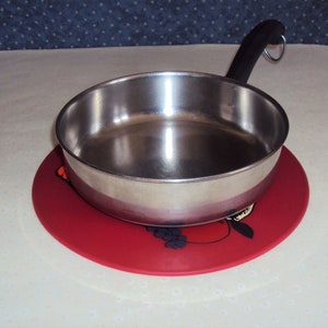 May include: A stainless steel saucepan with a black handle sits on a red silicone hot pad with a fruit design.