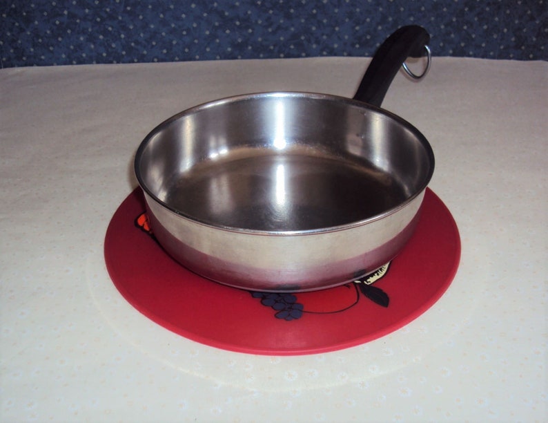 May include: A stainless steel saucepan with a black handle sits on a red silicone hot pad with a fruit design.