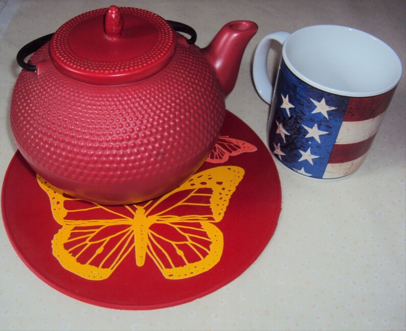 May include: Red teapot with a lid and a handle, sitting on a red coaster with a yellow butterfly design. A white mug with a red, white, and blue American flag design is next to the teapot.