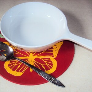 May include: A white ceramic bowl with a handle sits on a red trivet with a yellow butterfly design. A silver slotted spoon is on the trivet.