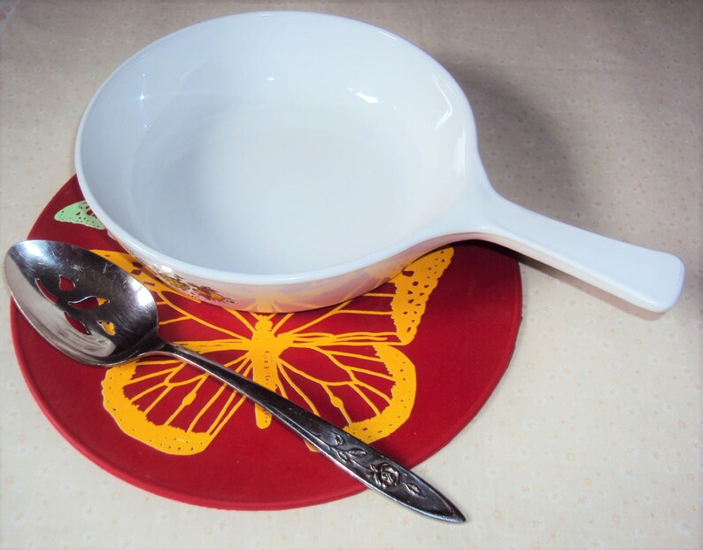 May include: A white ceramic bowl with a handle sits on a red trivet with a yellow butterfly design. A silver slotted spoon is on the trivet.