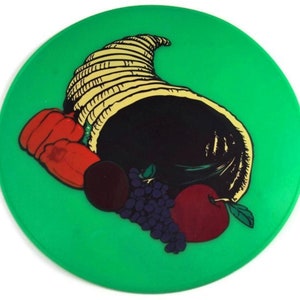 May include: A green round plate with a cornucopia filled with fruit and grapes. The cornucopia is brown and yellow with a black interior. The fruit includes a red apple and a red pepper.