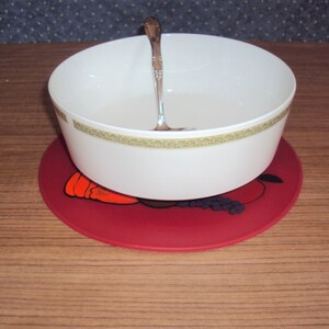 May include: A white ceramic bowl with a gold trim sits on a red silicone trivet with a fruit design. A silver spoon is inside the bowl.
