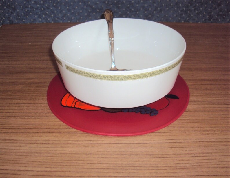 May include: A white ceramic bowl with a gold trim sits on a red silicone trivet with a fruit design. A silver spoon is inside the bowl.
