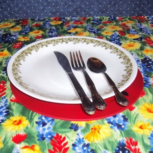 May include: A white plate with a green and gold floral pattern sits on a red placemat. A fork, knife, and spoon are placed on the plate. The placemat is on a tablecloth with a floral pattern.
