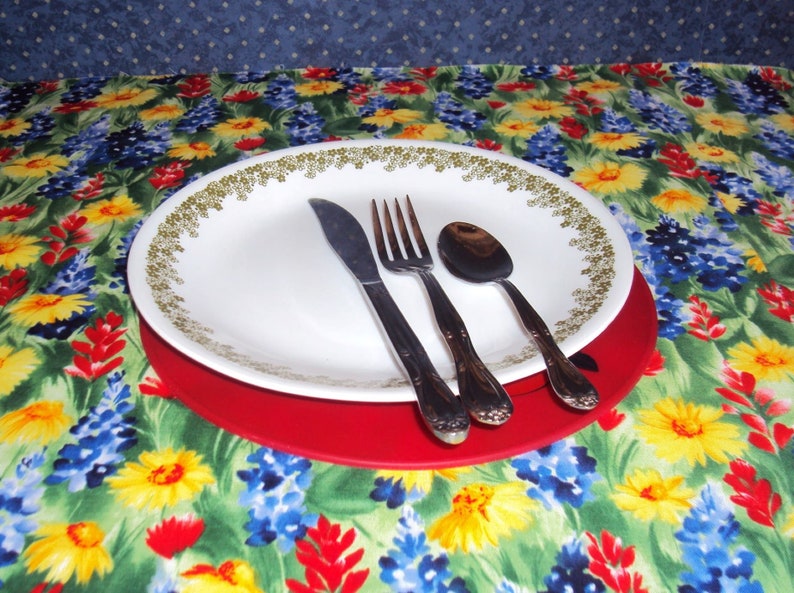May include: A white plate with a green and gold floral pattern sits on a red placemat. A fork, knife, and spoon are placed on the plate. The placemat is on a tablecloth with a floral pattern.