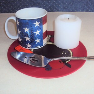 May include: A red round silicone trivet with a black and white illustration of a rooster and a silver serving spoon. A white candle and a mug with an American flag design are on the trivet.