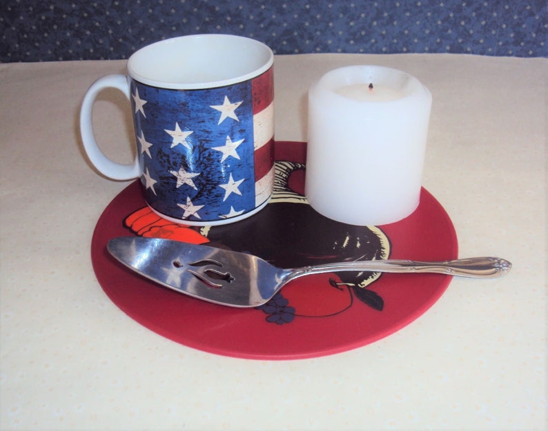 May include: A red round silicone trivet with a black and white illustration of a rooster and a silver serving spoon. A white candle and a mug with an American flag design are on the trivet.