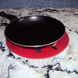 May include: A black frying pan with a black handle sits on a red and white patterned hot pad.