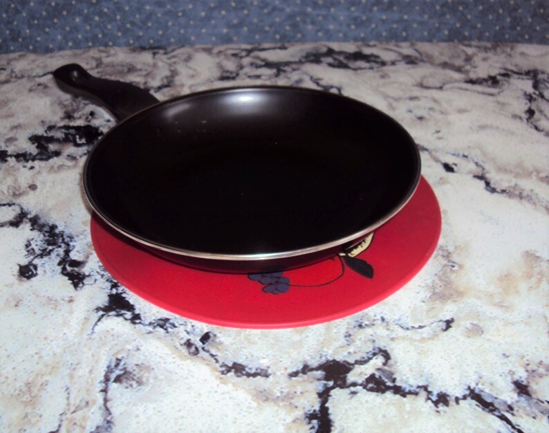May include: A black frying pan with a black handle sits on a red and white patterned hot pad.