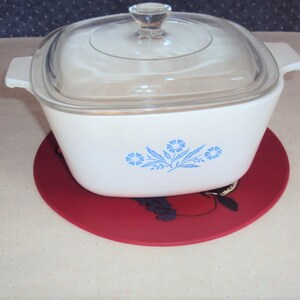 May include: White casserole dish with a blue floral design and a glass lid. The dish is sitting on a red trivet with a black grape design.