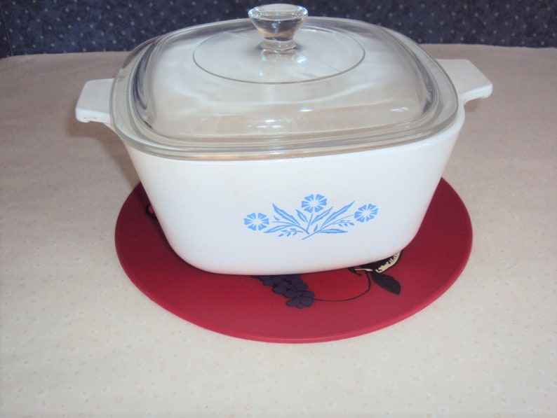 May include: White casserole dish with a blue floral design and a glass lid. The dish is sitting on a red trivet with a black grape design.