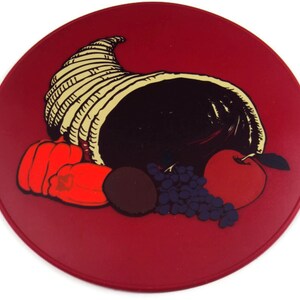May include: A red round plate with a cornucopia filled with fruit and vegetables. The cornucopia is made of woven straw and is filled with a red apple, red peppers, grapes, and a dark brown fruit.