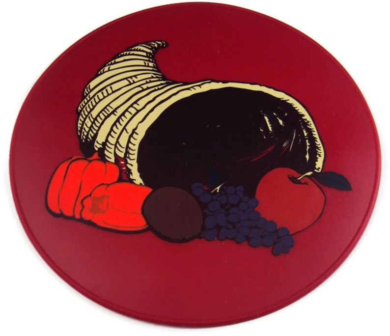 May include: A red round plate with a cornucopia filled with fruit and vegetables. The cornucopia is made of woven straw and is filled with a red apple, red peppers, grapes, and a dark brown fruit.