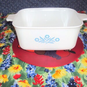 May include: White square baking dish with blue flower design on a red and black floral trivet.