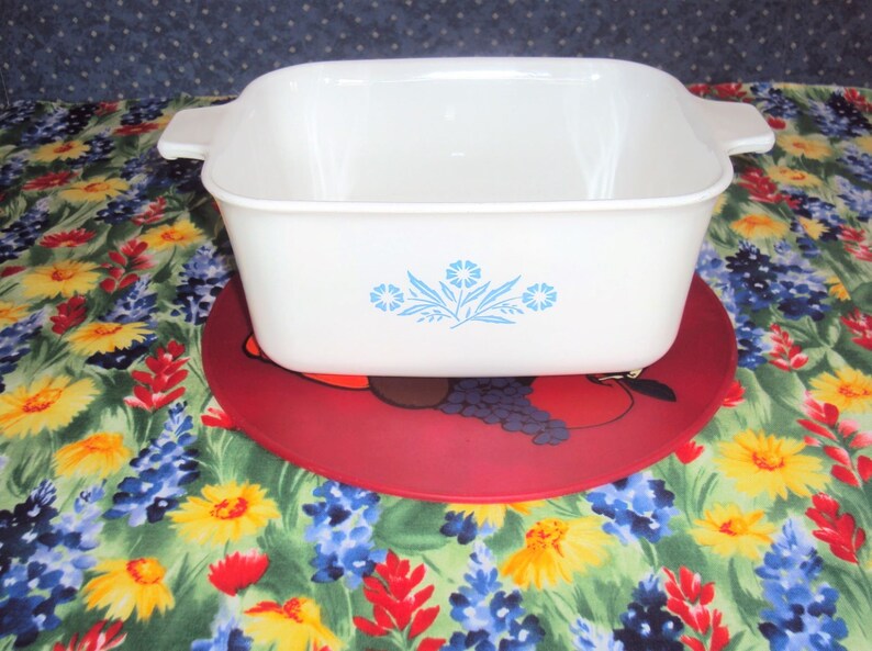 May include: White square baking dish with blue flower design on a red and black floral trivet.