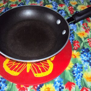 May include: A black non-stick frying pan with a black handle sits on a red trivet with a yellow butterfly design. The trivet is on a floral tablecloth with blue, yellow, and red flowers.