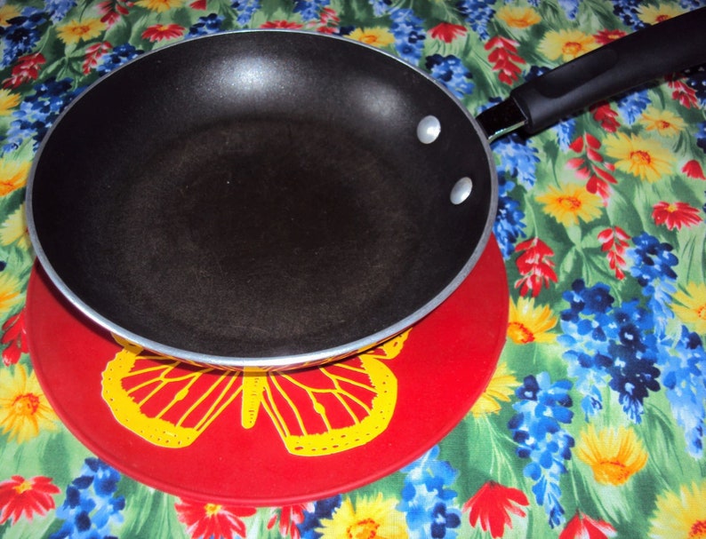 May include: A black non-stick frying pan with a black handle sits on a red trivet with a yellow butterfly design. The trivet is on a floral tablecloth with blue, yellow, and red flowers.