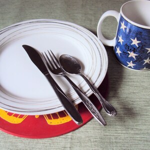 May include: A white plate with gold trim sits on a red placemat with a yellow design. A fork, knife, and spoon are placed on the plate. A white mug with a blue and white American flag design is in the background.