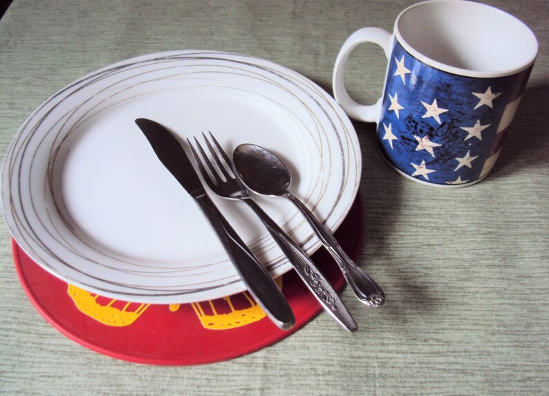 May include: A white plate with gold trim sits on a red placemat with a yellow design. A fork, knife, and spoon are placed on the plate. A white mug with a blue and white American flag design is in the background.