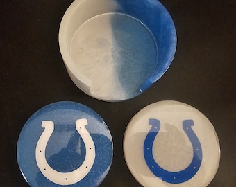May include: A round resin coaster holder with a split white and blue design, accompanied by two circular coasters. One coaster is blue with a white horseshoe logo, and the other is white with a blue horseshoe logo, both featuring a glossy finish.