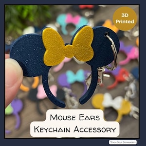 May include: A 3D-printed navy blue mouse ears keychain accessory with a gold bow. The keychain has a silver ring and clasp. The text "Mouse Ears Keychain Accessory" is displayed on the image.