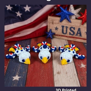 May include: Three 3D-printed eagle turtle figurines with patriotic colors. The figurines are white with blue, red, and yellow accents. The background includes a USA flag and a wooden sign that says "USA". The text reads "Patriotic Eagle Turtles".