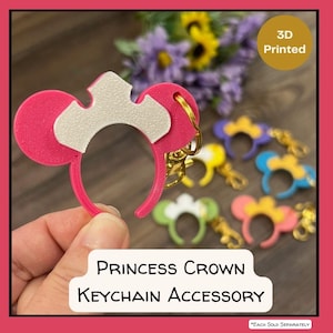 May include: A pink and white 3D-printed Princess Crown keychain accessory. The keychain features a pink headband with two circular ears and a white crown design. Other colorful keychains are visible in the background.