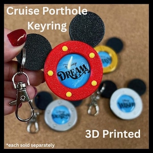 May include: A collection of 3D-printed cruise porthole keyrings. The keyrings feature a red, yellow, and blue design with the word "DREAM" in white. The keyrings have black mouse ears and a silver clasp. The text "Cruise Porthole Keyring" and "3D Printed" are also visible.