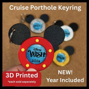 May include: A 3D printed keyring shaped like a mouse head with a red porthole frame featuring yellow accents and a central blue insert displaying the text Disney WISH 2026. Additional keyrings in white, gold, and silver are visible.