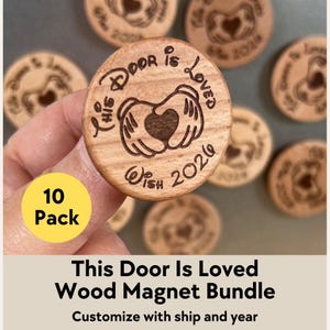 May include: A bundle of ten wooden magnets. Each magnet is round and features the text "This Door Is Loved Wish 2026" with a heart design. The magnets are held in a hand, with the text "10 Pack" in a yellow circle.