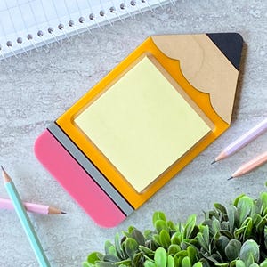 May include: A yellow and pink pencil-shaped sticky note holder with a yellow sticky note inside. The holder has a gray and black eraser on the top.