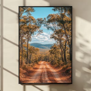 May include: A framed art print depicting a dirt track through a forest. The road is lined with tall trees with golden-brown leaves, leading to a distant mountain range under a blue sky with fluffy clouds. The artwork is encased in a black frame.