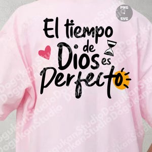 Peut inclure: T-shirt rose clair avec l'inscription espagnole "El tiempo de Dios es Perfecto" en script noir. Le design comprend un cœur rouge, un sablier et un soleil jaune.