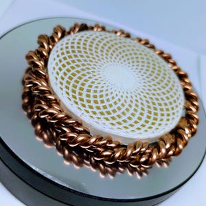 Könnte beinhalten: Ein weißes kreisförmiges Objekt mit einem geometrischen Muster, umgeben von einer dicken Kupferspule.