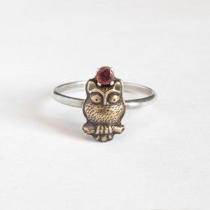 Tiny brass owl ring with a garnet above the head and a thin silver band. white background.